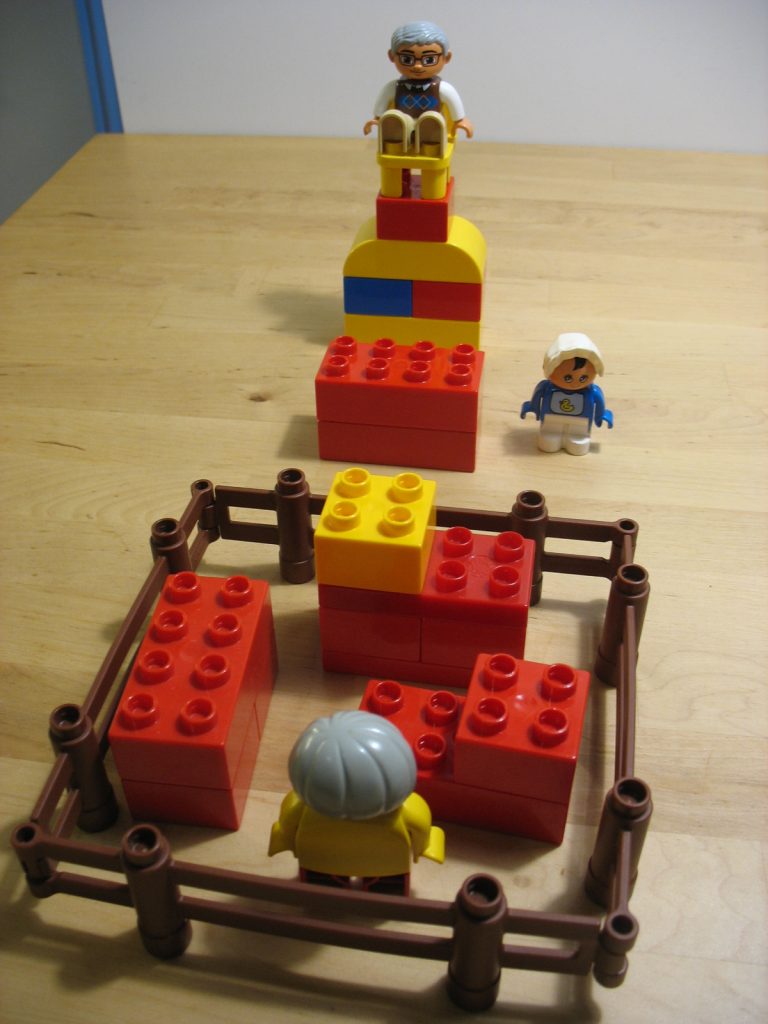 A scene of a play with Duplos. A character sitting up on a throne-looking high chair, a second character standing next to the throne and a third character standing in an enclosure with three blocks made out of a few Duplos.