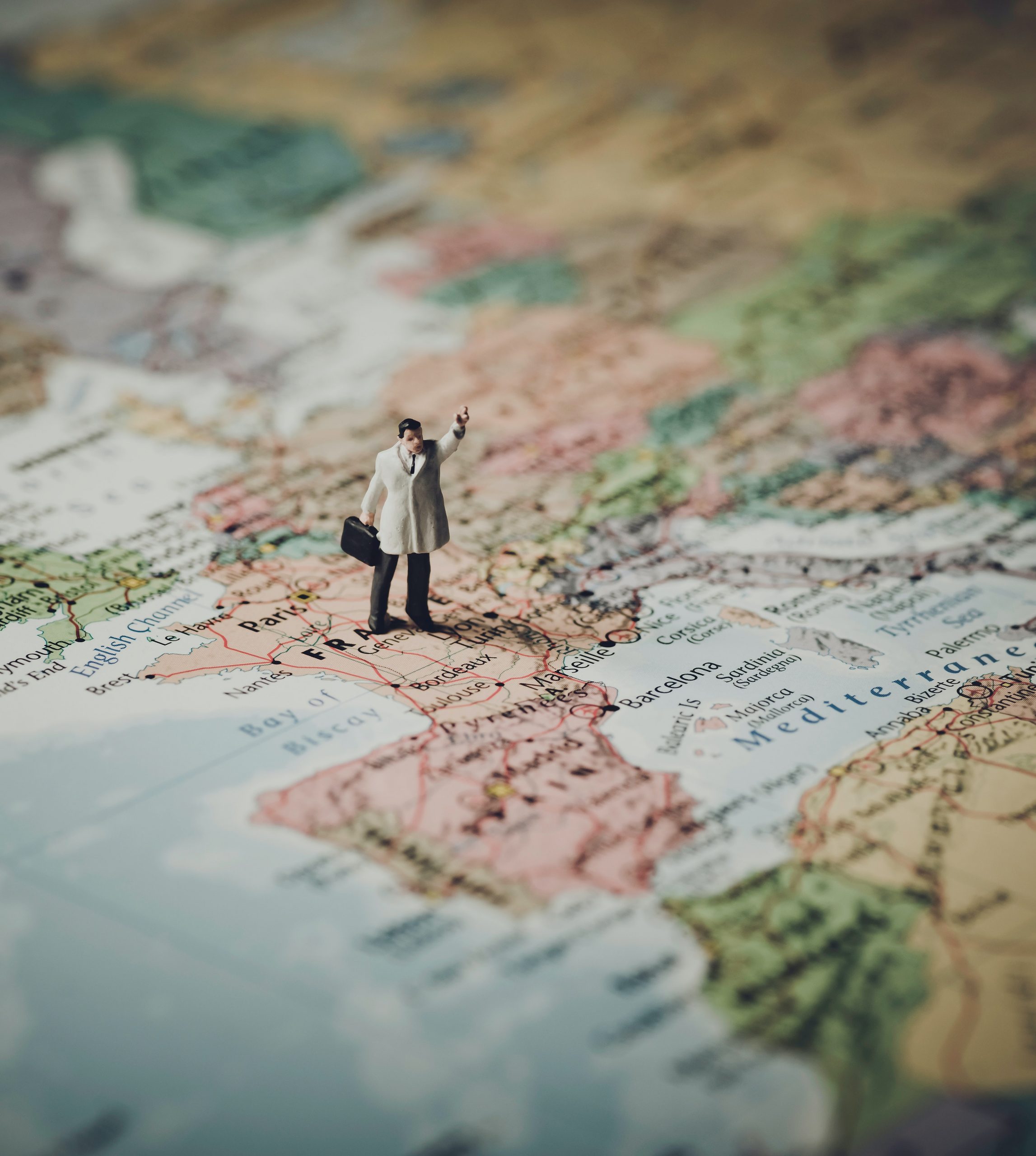 A closeup of a maps displaying a part of Europe and Africa. A small plastic male figure standing in the middle of France in a white coat, holding his left hand up and a briefcase in the left hand.